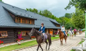Grupa osób na koniach przejeżdża obok drewnianego budynku stadniny.