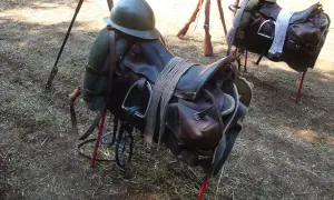 Dwa siodła, część rzędu końskiego, z hełmami i karabinami w tle.