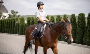 Młoda kobieta w kasku i rękawiczkach jedzie na kasztanowym koniu. Wygląda, jakby właśnie zdała egzamin na prawo jazdy na konia.
