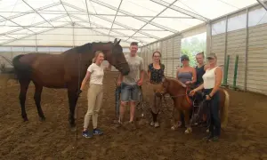 Grupa uczestników kursu jazdy konnej z instruktorem pozuje do zdjęcia z dwoma końmi, w tym kucykiem.