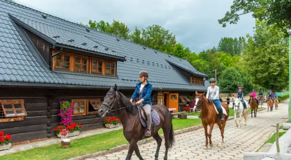 Grupa osób na koniach przejeżdża obok drewnianego budynku stadniny.